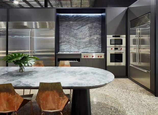 A dramatic contemporary kitchen and dining area with extensive stainless-steel appliances and cabinetry, a large illuminated veined stone feature splashback behind the cooktop, integrated ovens, and a broad oval stone island with a dark pedestal base. Warm-toned sculptural dining chairs, a tropical plant, polished terrazzo-like flooring, and an exposed industrial ceiling add contrast.
