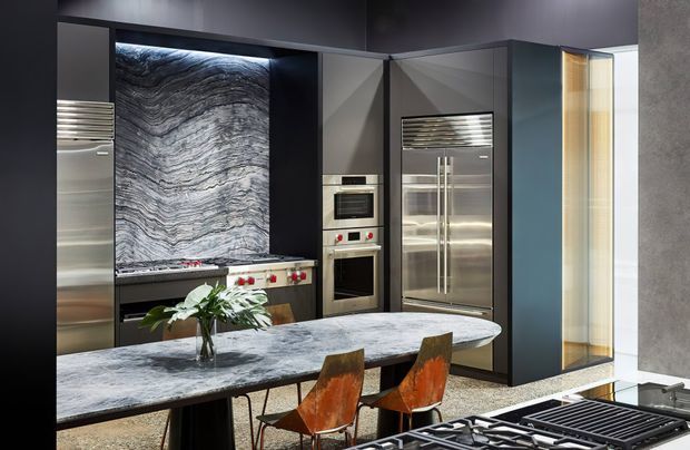 A luxurious contemporary kitchen with dark cabinetry, integrated stainless-steel appliances, a dramatic veined stone feature backsplash, and a large waterfall-edge island with dining seating. Warm timber accents and concealed lighting add contrast to the predominantly dark, cool-toned space.