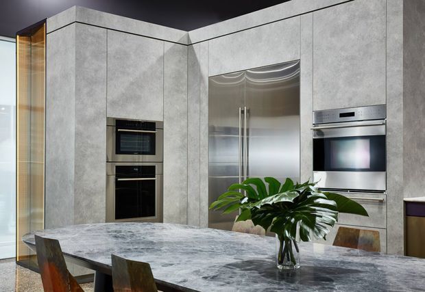 A sophisticated contemporary kitchen and dining area with full-height grey stone-look cabinetry, integrated black appliances, a stainless-steel double-door refrigerator, and a large grey marble-look island or dining table. Dark ceilings and timber-framed glazing create contrast, while a large green plant adds a natural accent.