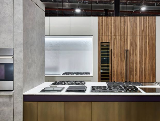 Contemporary commercial-style kitchen featuring a large island with multiple gas cooktops and integrated appliances. The kitchen combines white stone surfaces, warm timber cabinetry, grey concrete-look wall panels, and a partially exposed services ceiling with black track lighting.