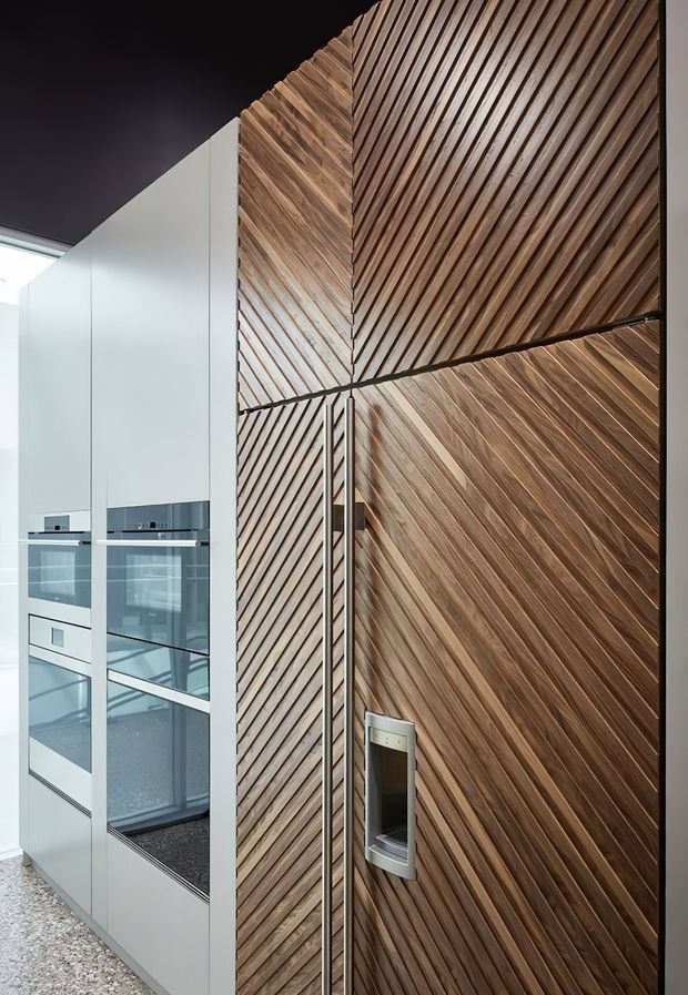 A close-up interior view of a contemporary kitchen featuring tall white built-in cabinetry with integrated ovens and reflective dark glass fronts, adjacent to a dramatic floor-to-ceiling timber feature wall. The timber is arranged in repeated diagonal slatted panels with contrasting directional patterns at the corner and horizontal break. A dark ceiling and polished, refined finishes create a modern architectural character.