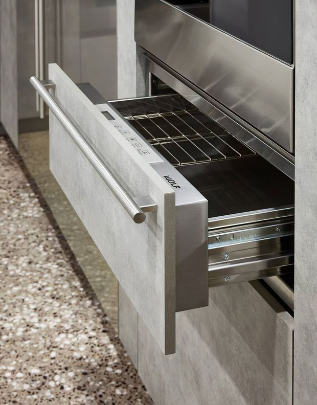 Close-up of a built-in stainless-steel oven with its door open, set into pale textured cabinetry or wall joinery. The oven has a large cylindrical metal handle, visible control panel, wire rack, and telescopic runners. A terrazzo-style floor is visible alongside the minimalist built-in installation.