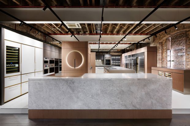 A spacious contemporary kitchen with a large pale stone island, warm timber cabinetry, integrated appliances and display shelving. The room combines a refined modern fit-out with exposed brick walls, exposed ceiling services and dark structural framing, creating an industrial character.