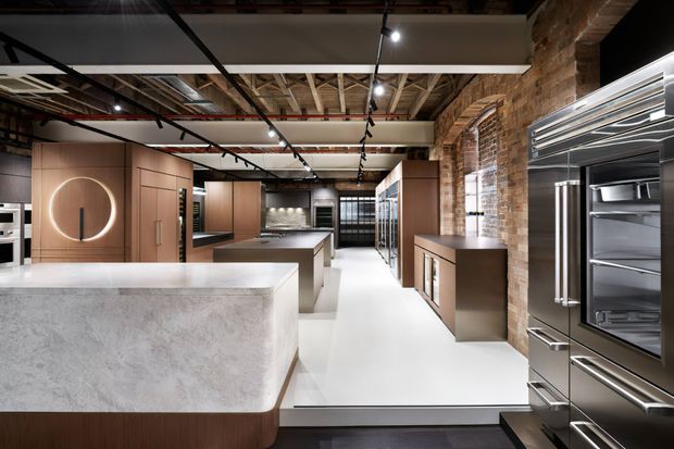 A sophisticated contemporary kitchen showroom or commercial kitchen display featuring a large pale stone island, dark timber cabinetry, integrated appliances, and extensive stainless-steel refrigeration. Exposed brick walls, exposed timber roof structure, black ceiling services, and track lighting create an industrial architectural character, while warm wood cabinetry and clean white surfaces keep the space refined and modern.