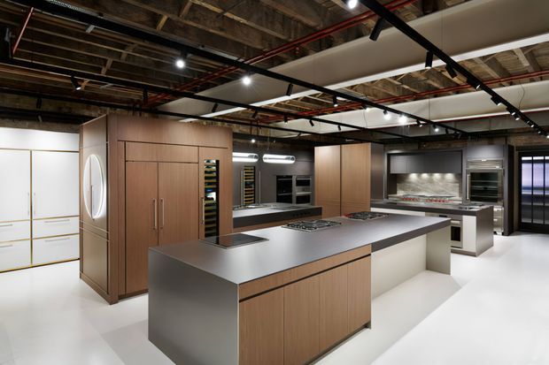 A large contemporary commercial-style kitchen with a central island, extensive integrated cabinetry, stainless-steel work surfaces, multiple cooking zones, built-in ovens, refrigeration and wine storage. The open ceiling exposes timber framing, services and red pipework, while suspended linear lighting and black track elements add an industrial character. The palette combines warm timber cabinetry, dark grey surfaces, white flooring and stainless-steel appliances.