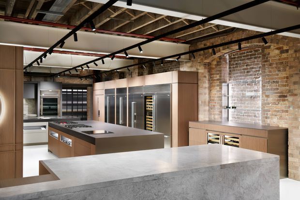 A sophisticated open-plan kitchen with a large concrete-look island and extensive timber cabinetry. Stainless-steel appliances, integrated wine storage, exposed brick walls, and an exposed structural ceiling are accented by black track lighting and visible services, creating an industrial yet refined commercial-style interior.