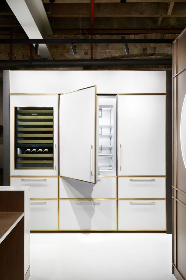 A built-in white cabinetry wall with slim brass-gold trim and long brass handles, featuring an integrated wine cooler and open refrigerator compartment. The cabinetry is set against a raw exposed brick and timber ceiling with visible black services and track lighting, creating a refined industrial kitchen or hospitality fit-out.