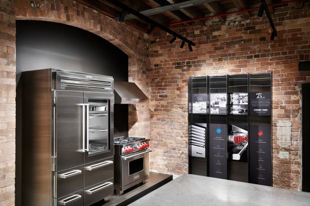 A sophisticated industrial-style kitchen showroom or display space with exposed red brick walls, a large arched brick opening, stainless-steel refrigerator and range cooker, dark cabinetry, a grey benchtop, and black track lighting. A tall black display or storage wall is visible on the right.