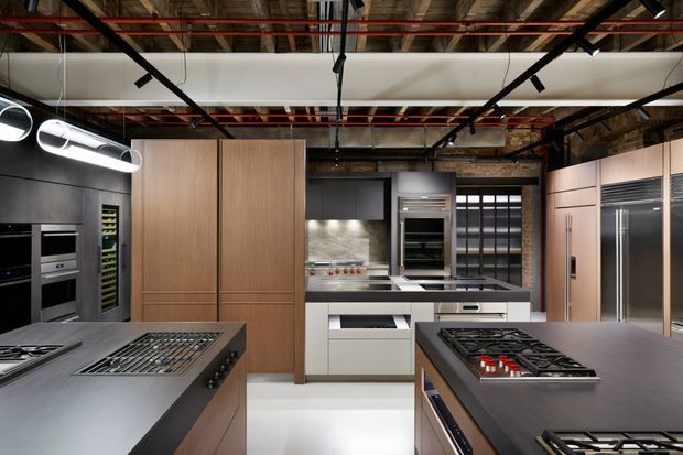 A large contemporary commercial-style kitchen with multiple cooking islands, integrated ovens, refrigeration and extensive timber-toned cabinetry. The space features dark grey benchtops, stainless-steel appliances, a concealed or exposed industrial ceiling with black track lighting and visible services, and a partially exposed brick wall at the rear.