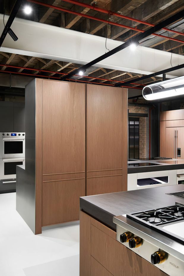 A contemporary kitchen with extensive warm wood cabinetry, tall integrated storage, a large dark benchtop island with gas cooktop, built-in ovens, and a secondary run of cabinetry. The space combines a crisp white floor and cabinetry with exposed timber ceiling structure, visible red services, black framing, and industrial-style linear lighting and ductwork.