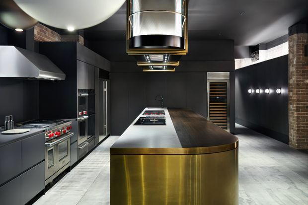 A dramatic, high-end contemporary kitchen with a long central island featuring a brushed brass/gold base, dark timber-toned benchtop, and integrated cooktop. Matte-black cabinetry, stainless-steel appliances, a sculptural rangehood, exposed brick accents, pale stone-look flooring, and oversized metallic pendant lighting create a bold industrial-luxe atmosphere.