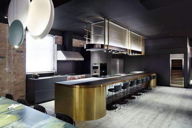 A dramatic high-end industrial-style kitchen with a large curved island clad in brushed brass, dark cabinetry, integrated stainless-steel appliances, a long breakfast bar with black stools, exposed brick, and a striking sculptural pendant light. A mezzanine-like overhead structure with brass railings adds architectural interest, while pale stone or concrete-look flooring contrasts with the dark ceiling and joinery.