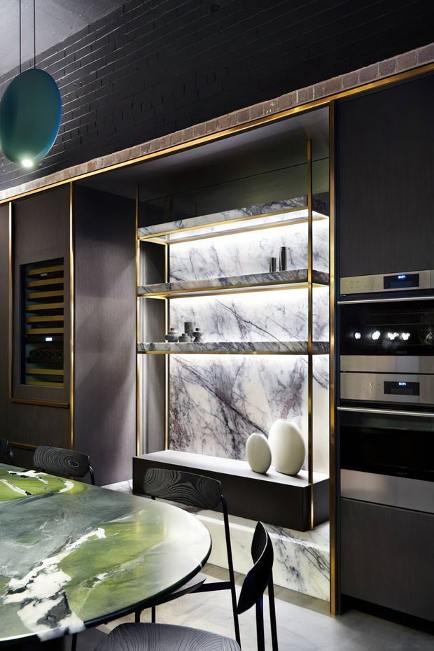 A dramatic, high-end kitchen and dining area with dark cabinetry, integrated stainless-steel ovens, wine storage, illuminated open shelving, and a striking veined marble feature wall. A round green-and-white stone dining table with dark sculptural chairs occupies the foreground. The space has a black painted brick ceiling and wall, warm linear lighting, and brass-toned trim details.