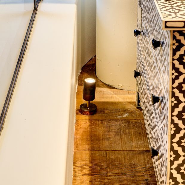 Close-up interior detail showing a narrow passage beside a white bathtub or large fixture, a warm-toned timber floor, and a patterned built-in cabinet with dark knobs. A small cylindrical accent lamp sits on the floor, creating a warm ambient glow.