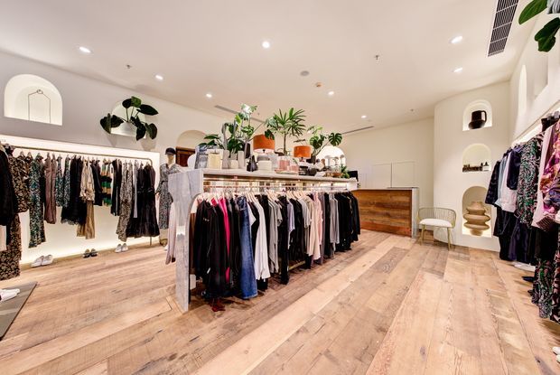 Bright boutique clothing shop with clothing racks arranged across a spacious retail interior. The room features pale timber-look flooring, white plaster walls with rounded arched niches, recessed lighting, a central display island with potted plants and folded merchandise, and a timber-fronted counter or cabinet at the rear.