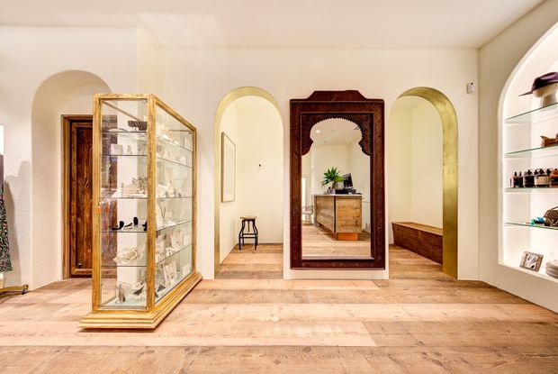 Warm, refined retail or dressing-room interior with pale timber flooring, cream walls, several arched openings, built-in curved display shelving, a glass-and-gold display cabinet, a dark carved timber-framed mirror, and a timber console. The space has an eclectic, vintage-inspired character with strong symmetry and decorative detailing.