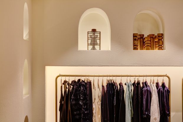 A warm, softly lit boutique clothing display set within a textured plaster wall. An open brass-toned rail holds a curated selection of garments, while recessed arched niches above display small decorative objects and folded textile or woven items. The space has an intimate, refined retail aesthetic with rounded architectural detailing.