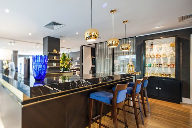 A sophisticated open-plan kitchen and home bar area featuring a long black marble island with brass edging, dark timber cabinetry, blue upholstered bar stools, and multiple gold globe pendant lights. A glass-fronted drinks display cabinet and illuminated shelving form a prominent feature wall, while a large blue vase and floral arrangement add colour. The space has a polished, luxurious contemporary interior with warm timber flooring and dark finishes.