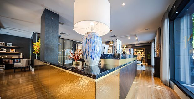 A sophisticated contemporary open-plan interior centred on a sculptural reception or kitchen island with a textured gold-toned base, dark stone benchtop, and a large decorative glass pendant light. Warm timber flooring, dark feature walls, extensive glazing, display shelving, and refined furnishings create a luxurious hospitality or residential entertaining atmosphere.