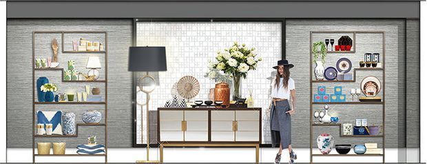 A stylised interior display vignette featuring symmetrical open shelving filled with decorative ceramics, books and objects, a central white sideboard with brass-toned hardware, a large floral arrangement, a table lamp, framed artwork and a person standing beside the console. The space has a dark grey textured wall finish, pale geometric feature tiling behind the centre, and a highly curated showroom-like composition.