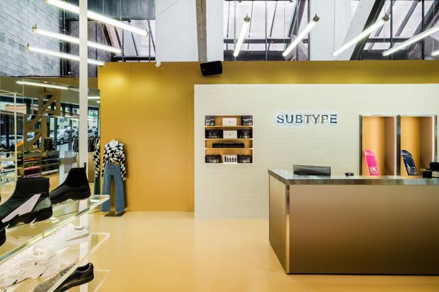 A contemporary retail shoe store interior with a prominent reception or checkout counter, illuminated product shelving, display tables, mannequins, and a branded wall sign reading “SUBTYPE.” The space features mustard-yellow painted walls and flooring, a white tiled feature wall, metallic counter finishes, exposed ceiling services, and linear pendant lighting.
