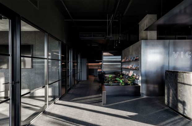 A dark, sophisticated commercial interior with a long entry corridor, black-framed glass partitions on the left, polished dark flooring, exposed black ceiling services, and a metallic reception or feature wall on the right. Shelving with brochures or products and built-in planter boxes line the space, while strong daylight casts geometric shadows across the floor.