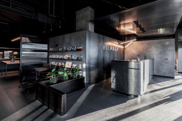 A dark, industrial-style commercial reception and waiting area with a black stone reception desk, charcoal concrete-look walls, black ceiling services, integrated shelving displaying brochures, office chairs, and indoor plants. Strong sunlight enters from the right, creating dramatic diagonal shadows across the floor. The space is branded “VICPROP” and appears to be a real-estate or professional office lobby.
