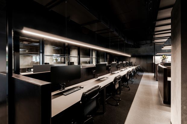A dark, sophisticated open-plan office interior with a long shared workstation and numerous computer monitors and office chairs. Black and charcoal finishes dominate, with exposed dark ceiling services, linear integrated lighting, glazed partitions, and a concrete-toned floor. The space has an industrial, dramatic character and appears to extend toward a brighter kitchenette or breakout area at the rear.