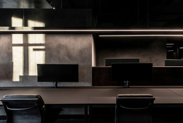 A dark, sophisticated shared workspace with a long built-in desk, multiple computer monitors, black office chairs, and a continuous linear light beneath a dark ceiling. The rear wall has a textured grey finish, while strong window light creates geometric shadows across it. The space has a refined industrial office character with restrained materials and subdued tones.