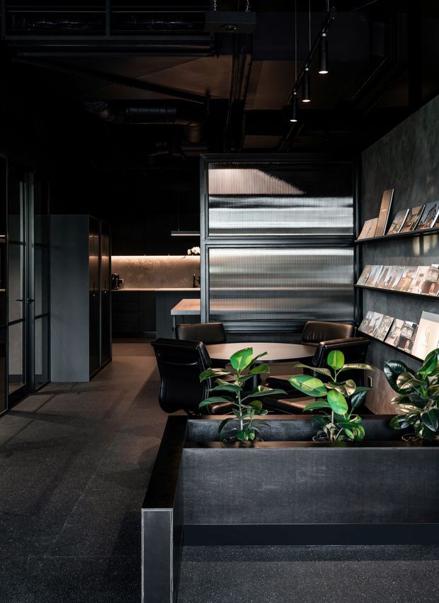 A dark, sophisticated commercial-style interior with an open-plan meeting or lounge area. Black-painted exposed services and ceiling structure create an industrial atmosphere, while a ribbed translucent partition, dark built-in shelving displaying printed materials, a compact kitchen or bar area, leather lounge chairs, and large integrated planter boxes define the space. The palette is predominantly black, charcoal, and dark grey with warm linear lighting and green indoor plants providing contrast.