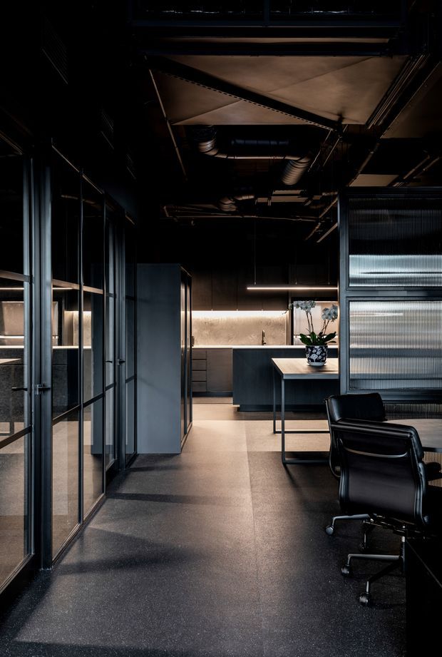 A dark, sophisticated contemporary office interior with exposed ceiling services, black-framed glazed partitions, a central worktable, black executive chairs, and a compact kitchenette or utility joinery area at the rear. Ribbed translucent glazing, warm linear lighting, dark flooring, and charcoal cabinetry create an industrial, refined atmosphere.