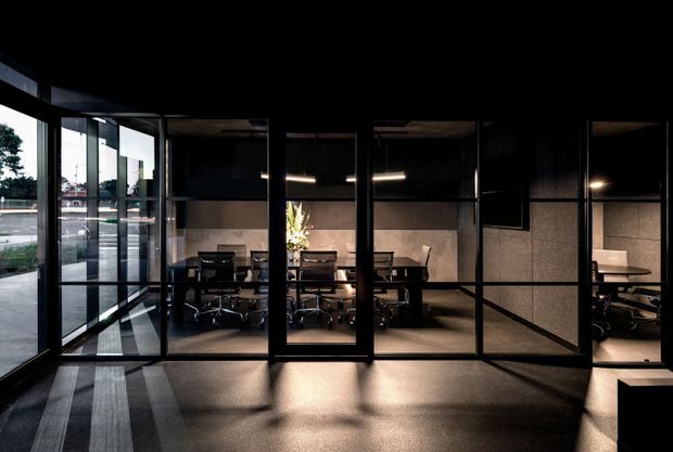 A dark, contemporary commercial interior viewed through extensive black-framed glass partitions. Several meeting or conference rooms contain desks and office chairs, with warm linear lighting, exposed dark ceilings, and neutral textured wall finishes. Strong reflections and daylight enter from the glazed facade on the left.