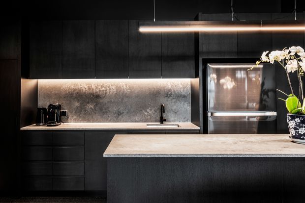 A dramatic contemporary kitchen in a dark palette, featuring matte black full-height cabinetry, a large central island with a pale stone benchtop, a textured grey splashback, integrated stainless-steel appliances, and under-cabinet and linear pendant lighting. A sink and black tap sit beneath the cabinetry, with an orchid adding a small natural accent.