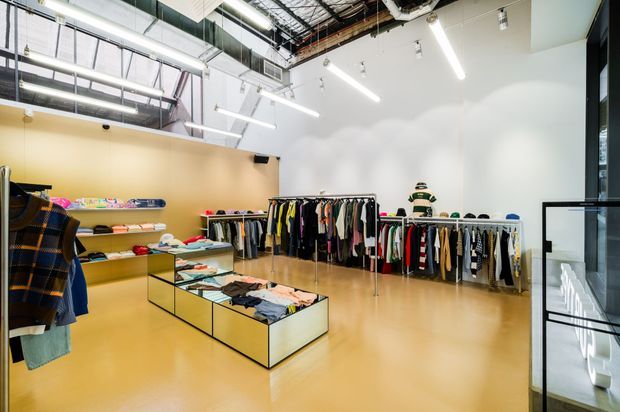 Bright modern clothing retail showroom with open garment racks, folded apparel on low glass display tables, wall shelving, mannequins, and a polished warm-toned floor. The space features white walls, exposed ceiling services and ductwork, black-framed glazing, and linear track lighting.