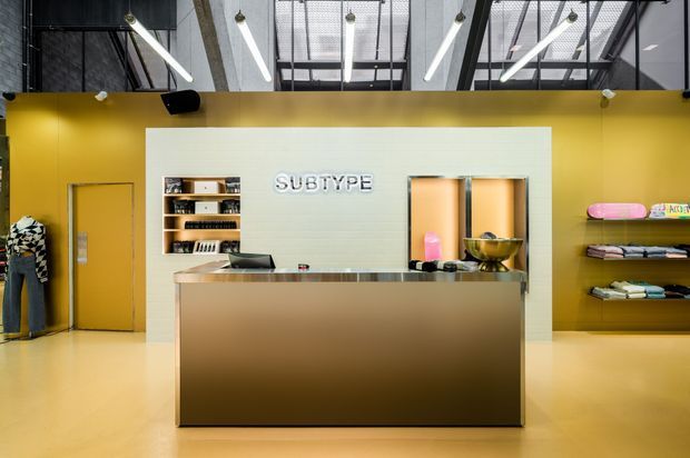 A bold contemporary retail reception area with a large metallic service counter centered in front of a pale ribbed feature wall bearing the word “SUBTYPE.” Mustard-yellow walls and flooring surround open shelving displaying merchandise, while exposed dark ceiling structure, extensive glazing, and linear pendant lighting create an industrial-style commercial interior.