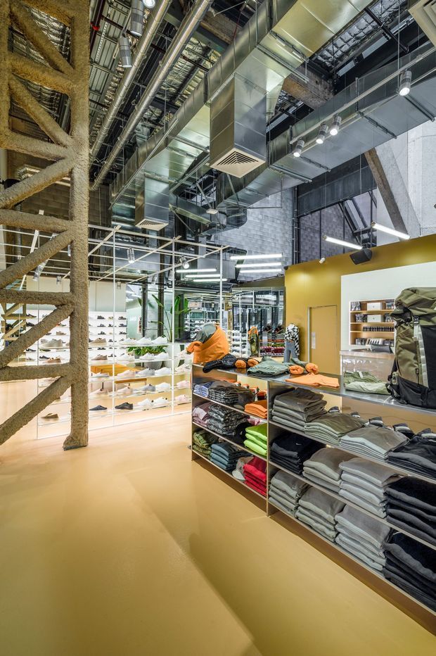 Interior view of a contemporary retail clothing and footwear store. The space features exposed industrial-style ceiling services and ductwork, a textured concrete block wall, suspended track and linear lighting, large mirrors, open shelving, and folded apparel displays. A prominent structural or decorative timber-like diagonal braced element frames the left side, while mustard-yellow painted walls and warm tan flooring add colour to the otherwise metallic industrial fit-out.