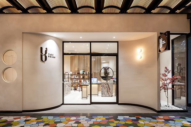 A contemporary commercial storefront entrance with a recessed glass-and-black-metal entry, curved plaster walls, warm interior lighting, and branded signage. The ceiling features a distinctive dark exposed grid with repeated illuminated slatted panels, while the foreground has colourful patterned paving and a side display window with a sculptural plant.