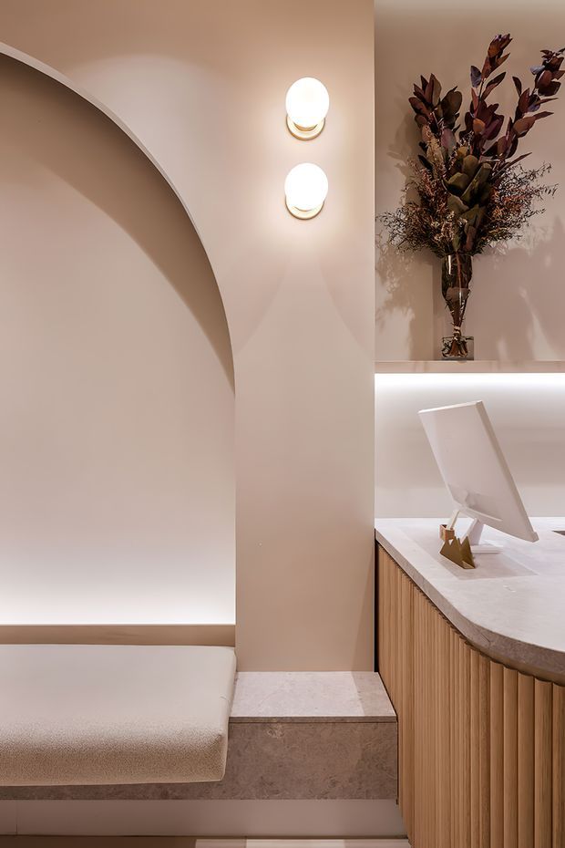 A refined, softly lit interior corner featuring a curved built-in upholstered bench beneath a large arched wall detail. Two oval wall sconces illuminate warm beige plaster walls. To the right is a curved reception or service counter with vertically fluted timber cabinetry, a pale stone benchtop, integrated display monitor, and a tall dried botanical arrangement in a wall niche. The overall design is quiet, sculptural, and luxurious.
