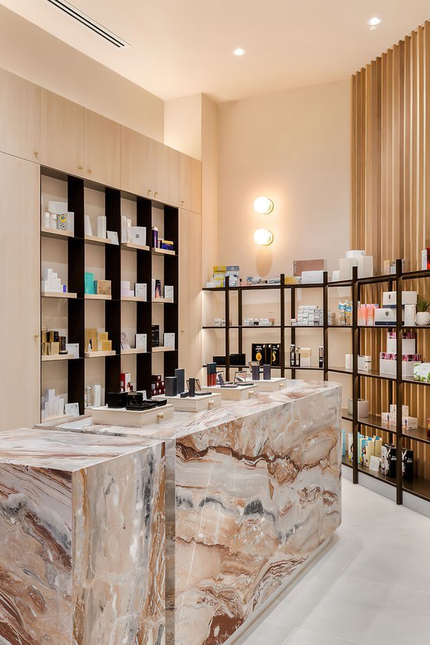 A refined retail interior, likely a beauty or skincare shop, with a large sculptural veined stone service counter in the foreground. Pale timber cabinetry with integrated display niches lines the left wall, while open black metal shelving displays products along the rear and right sides. Vertical timber screening, warm wall sconces, recessed ceiling lighting, and a light floor create a warm, luxurious and carefully curated atmosphere.