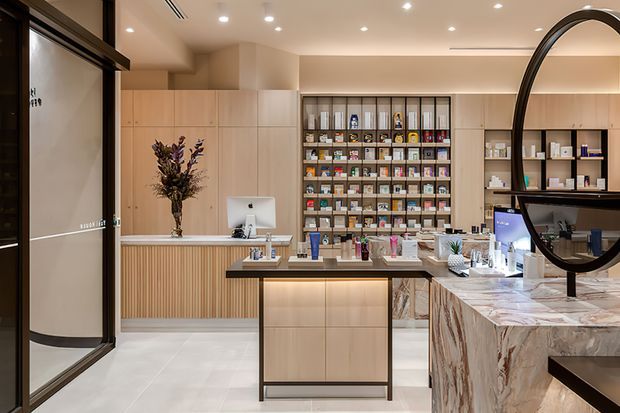 A refined retail pharmacy or wellness store interior with pale timber cabinetry, a central display island, product shelving, a ribbed timber reception counter, and a dramatic veined stone service counter in the foreground. Black-framed glazed entry doors and a large circular black-framed mirror add strong graphic accents. The space is brightly lit with recessed ceiling lights and concealed linear lighting.
