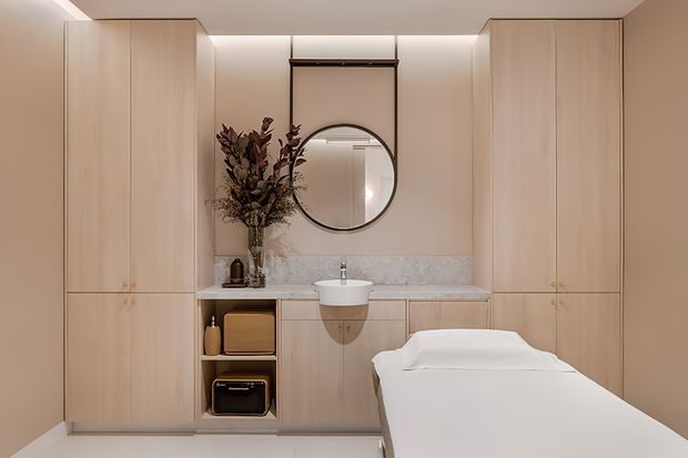 A softly lit, minimalist treatment room with a central massage table positioned in front of a built-in vanity and basin. Full-height pale timber cabinetry frames the space, with a marble-look splashback, round wall mirror, integrated concealed lighting, and open shelving containing small storage items. The neutral palette and dried botanical arrangement create a calm, refined atmosphere.