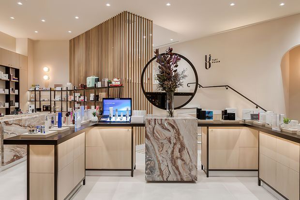 A refined contemporary retail beauty or wellness showroom with pale timber-toned display cabinetry, dramatic veined stone counters, open product shelving, integrated lighting, and a sculptural circular feature mirror with floral arrangement. A vertical timber-slat screen forms a strong architectural backdrop, while a stair and branded wall signage are visible behind the central display.