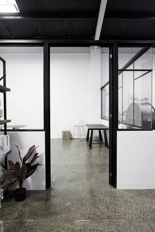 A minimalist industrial-style interior viewed through black-framed glazed partitions and an open glass door. The space features polished exposed aggregate/concrete flooring, white painted brick walls, black exposed ceiling structure, sparse black tables and stools, and a potted plant in the foreground. The glazing reveals an adjacent open workspace or studio area.