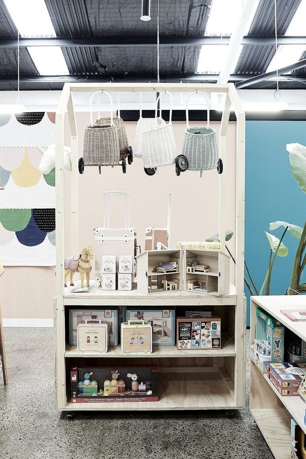 A bright retail display within an open industrial-style shop, featuring a pale timber freestanding shelving unit stocked with children's toys and small products. Three woven toy prams hang from the top frame, while colourful textiles and illustrated merchandise surround the display. The space has exposed dark corrugated roofing, white structural beams, polished concrete flooring, and a vivid blue wall with large indoor plants.