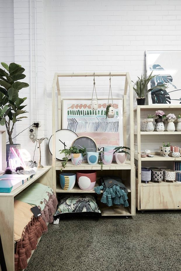 A bright, eclectic retail or studio-style interior with white-painted brick walls, polished speckled concrete flooring, pale timber shelving, and abundant colourful handmade ceramics, textiles, artwork, and indoor plants. A central open display unit features hanging artwork and decorative mugs, while a larger shelving unit on the right holds pottery and plants. The space has a playful, craft-oriented atmosphere with strong natural light.
