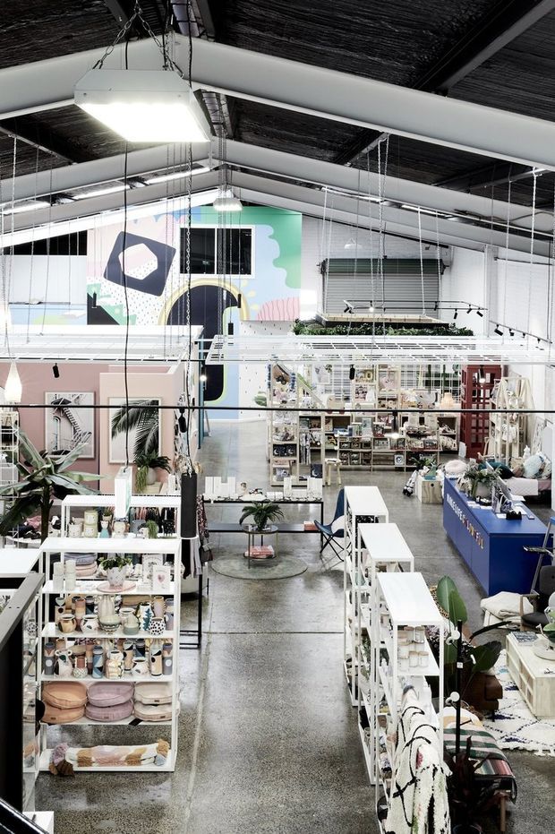 A bright, open-plan retail showroom with tall ceilings, exposed structural beams, suspended track and pendant lighting, polished concrete flooring, white display shelving, a blue service counter, and colourful merchandise arranged throughout. A large playful mural and mezzanine-like display structure add visual interest.