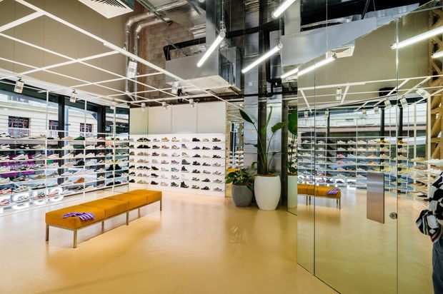 A bright contemporary shoe retail showroom with extensive wall-mounted and freestanding shoe displays, mirrored glazing, a central upholstered bench, polished light flooring, exposed services, and linear LED lighting. Large windows and reflective surfaces create an open, spacious commercial interior.