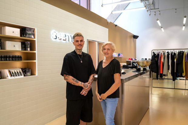 Bright contemporary retail interior with two people standing beside a polished reception or service counter. The space features white rectangular wall tiles, built-in timber display shelving with products, a glowing wall sign, clothing racks, and a high ceiling with exposed services and track lighting.