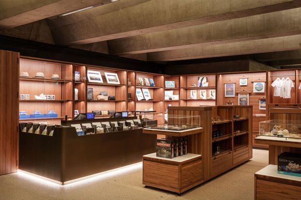 A warm, wood-panelled retail or exhibition showroom with built-in display shelving, glass-topped counters, framed prints, books, clothing, and small merchandise displays. The space has a dark ceiling with exposed structural beams, integrated shelf lighting, and timber display cabinetry arranged around a central circulation area.