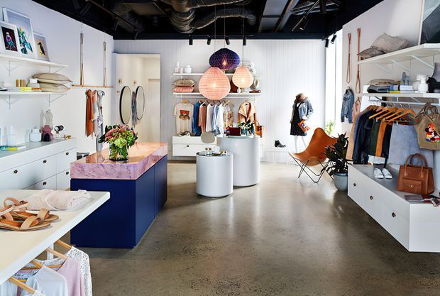 Bright contemporary retail showroom or boutique interior with white built-in shelving and display cabinetry, clothing racks, accessories, handbags, decorative objects, and central cylindrical display tables. The space has a polished concrete floor, white vertical wall cladding, a vivid blue-purple island display, colourful pendant lights, and an exposed black industrial ceiling with visible services.